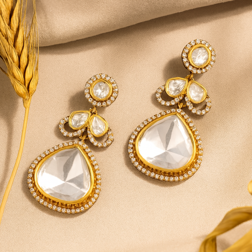 Gold and crystal earrings on a beige fabric background with dried wheat stalks.