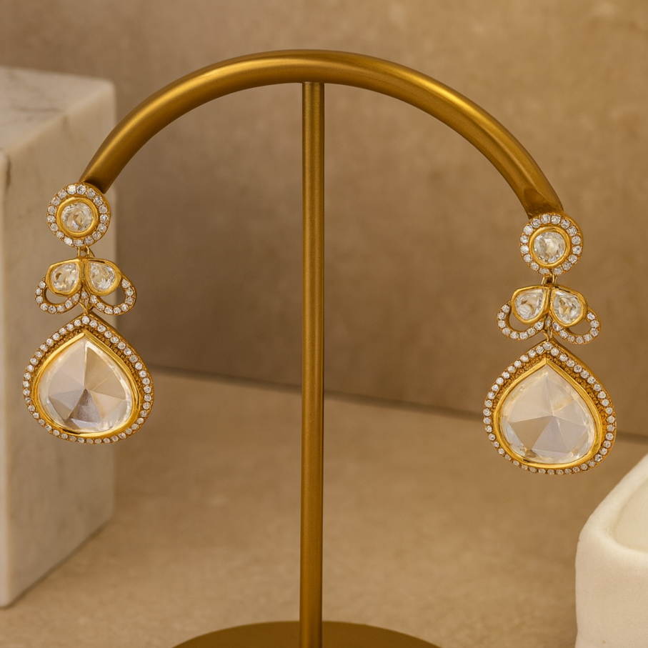 Gold earrings with teardrop-shaped designs on a gold stand against a beige background