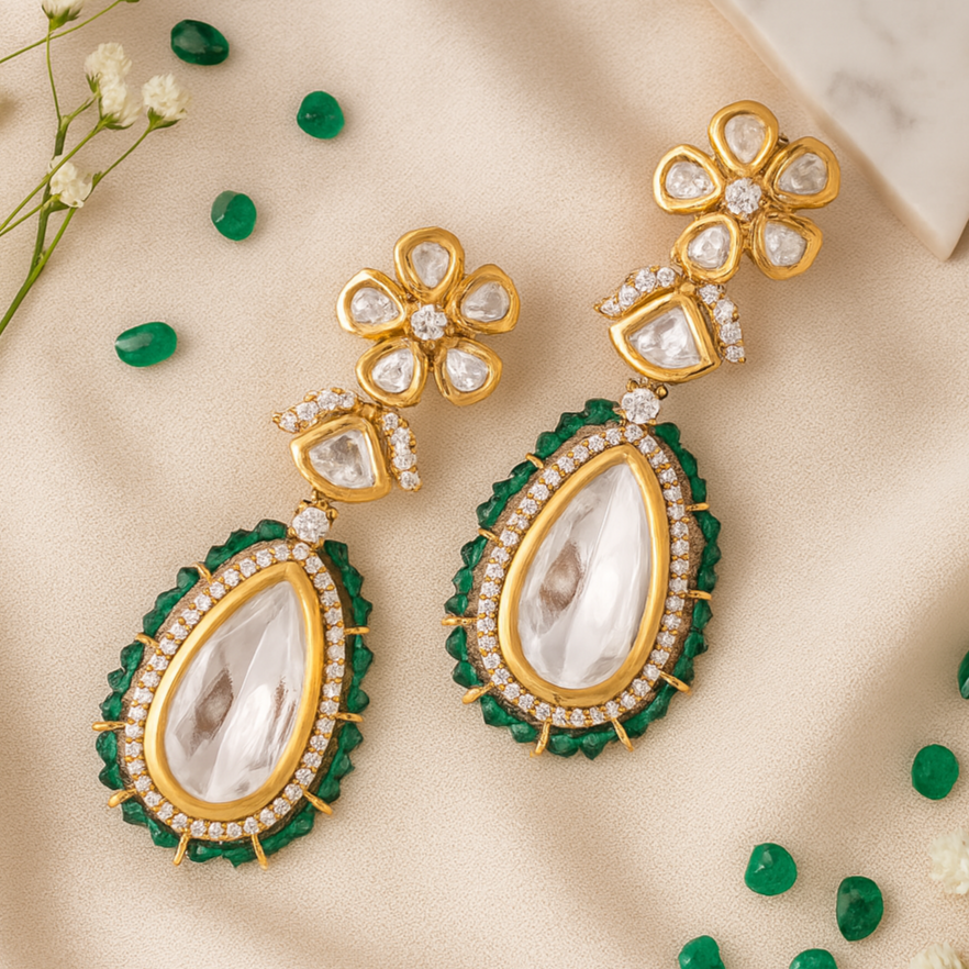 Gold and green earrings on a beige fabric background with small green stones and white flowers.