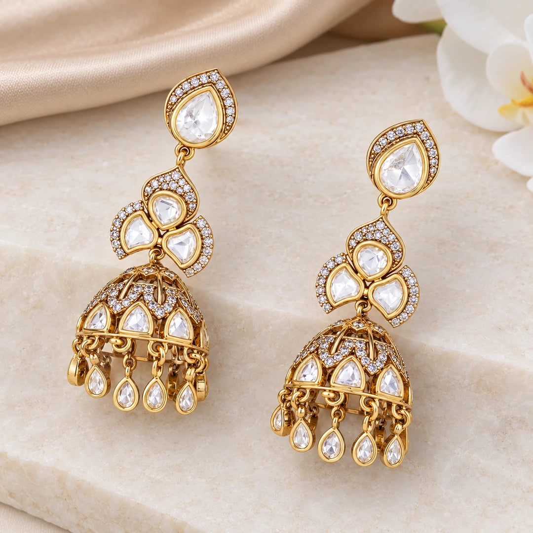 Pair of Kashika clear and gold-tone jhumka earrings on cream stone beside champagne silk and a white orchid.