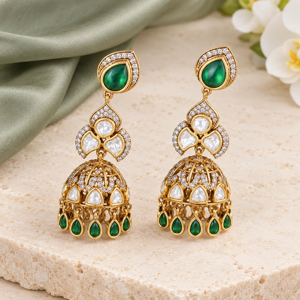 Pair of Kashika green and gold-tone jhumka earrings on cream stone beside sage silk and a white orchid.