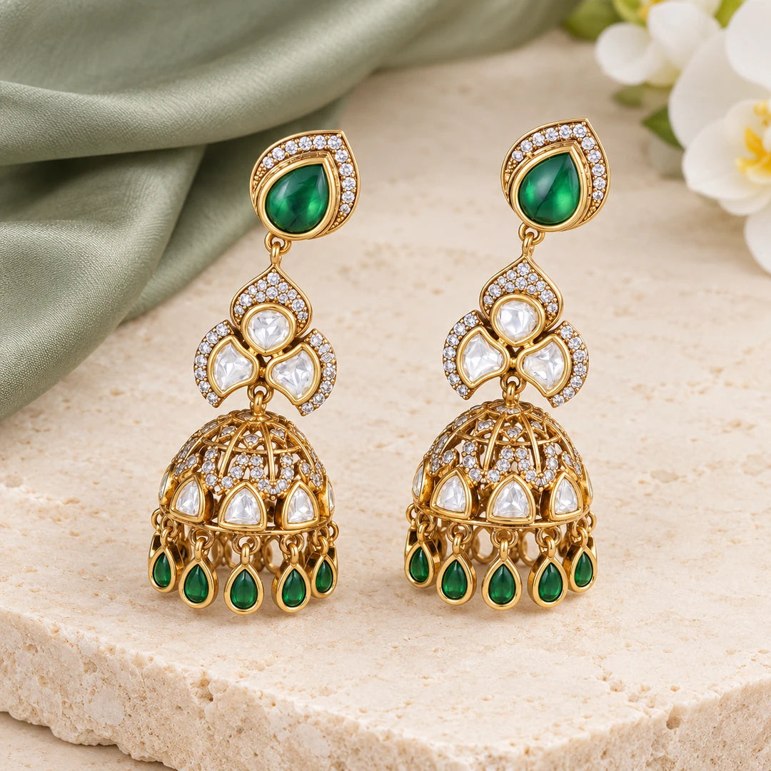 Pair of Kashika green and gold-tone jhumka earrings on cream stone beside sage silk and a white orchid.