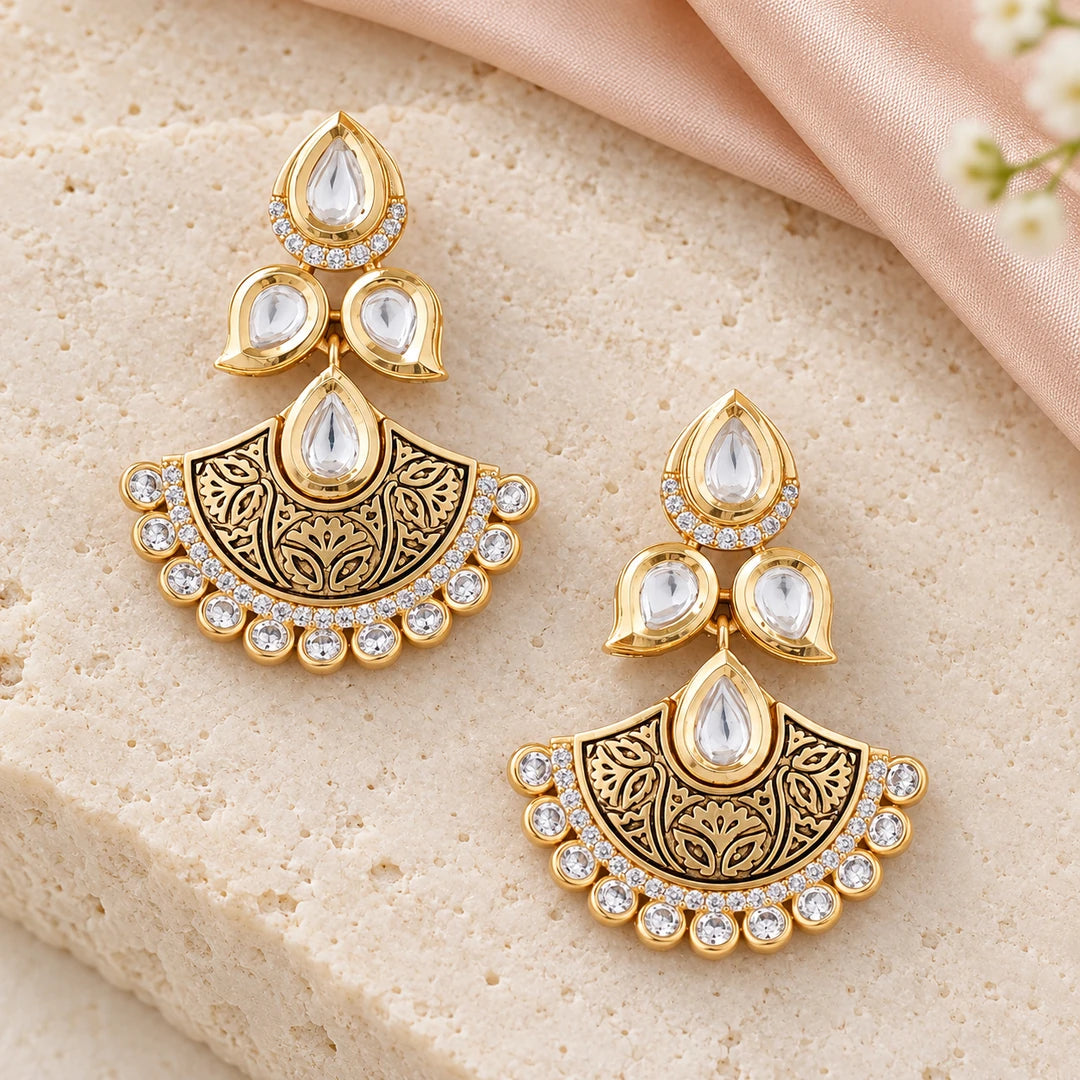 Pair of Kavya gold-tone floral chandbali earrings on pale cream stone beside blush silk and small white flowers.