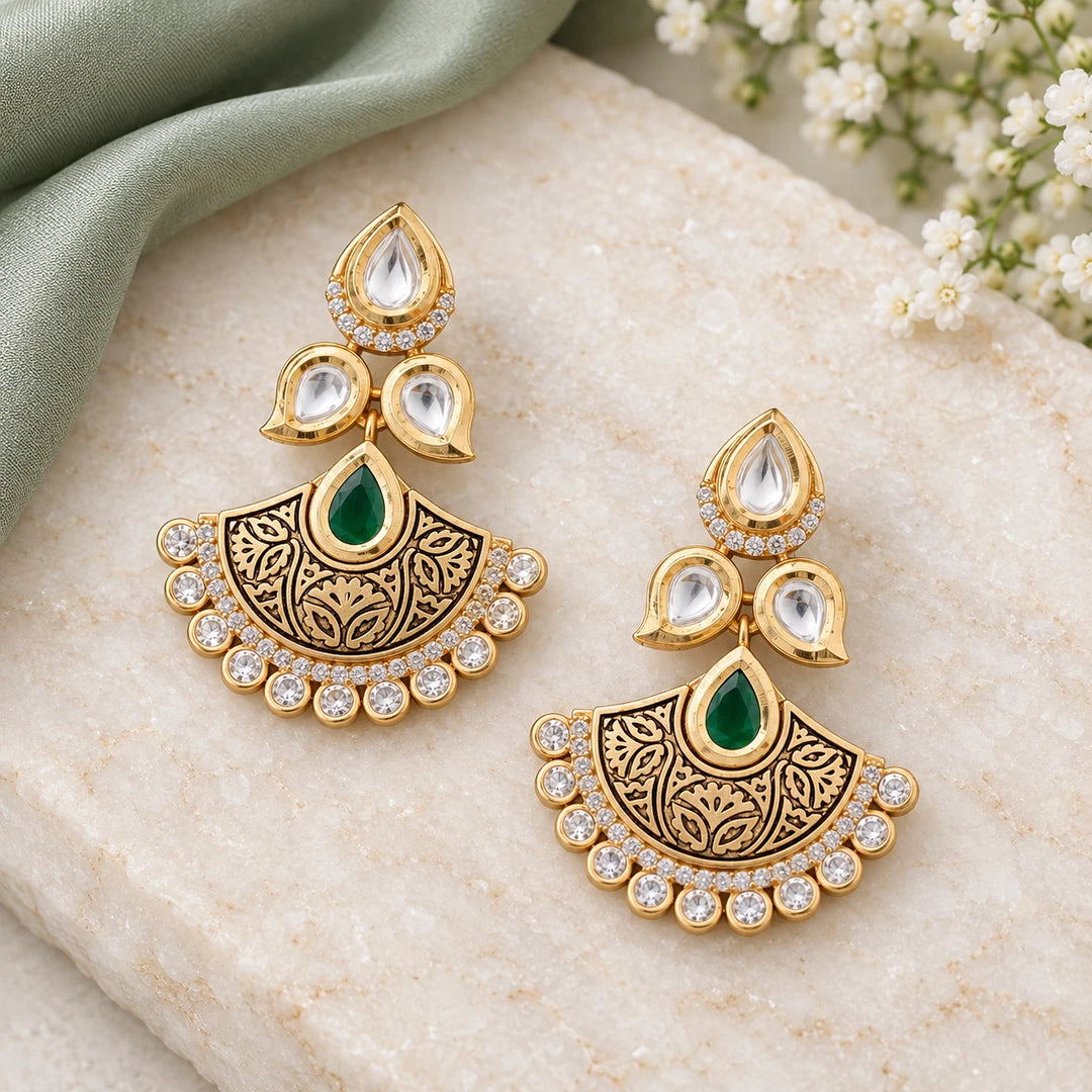 Pair of Kavya green and gold-tone floral chandbali earrings on cream stone beside sage silk and small white flowers.