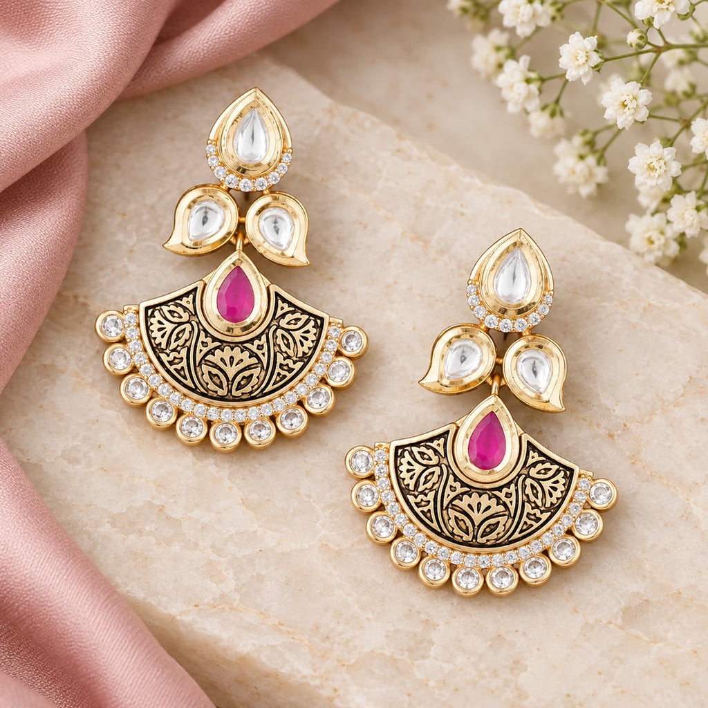 Pair of Kavya ruby-colour floral chandbali earrings on cream stone beside dusty rose silk and small white flowers.
