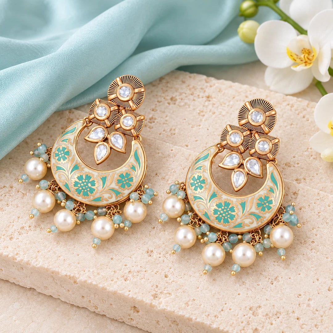 Pair of Kumud ivory and aqua floral chandbali earrings on cream stone beside pale aqua silk and a white orchid.