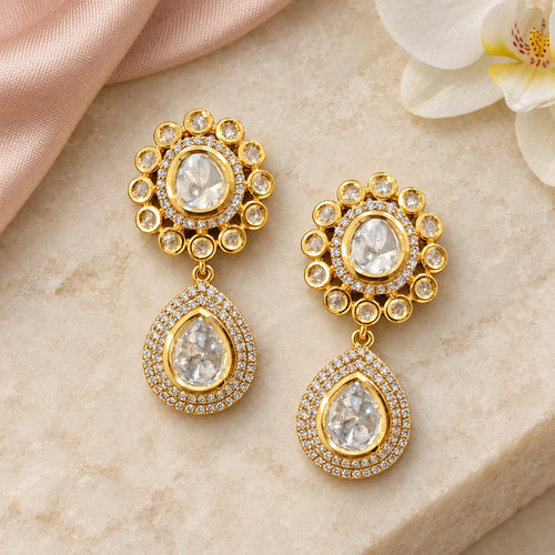 Pair of Aabha clear stone teardrop earrings on ivory stone beside blush silk and a white orchid.