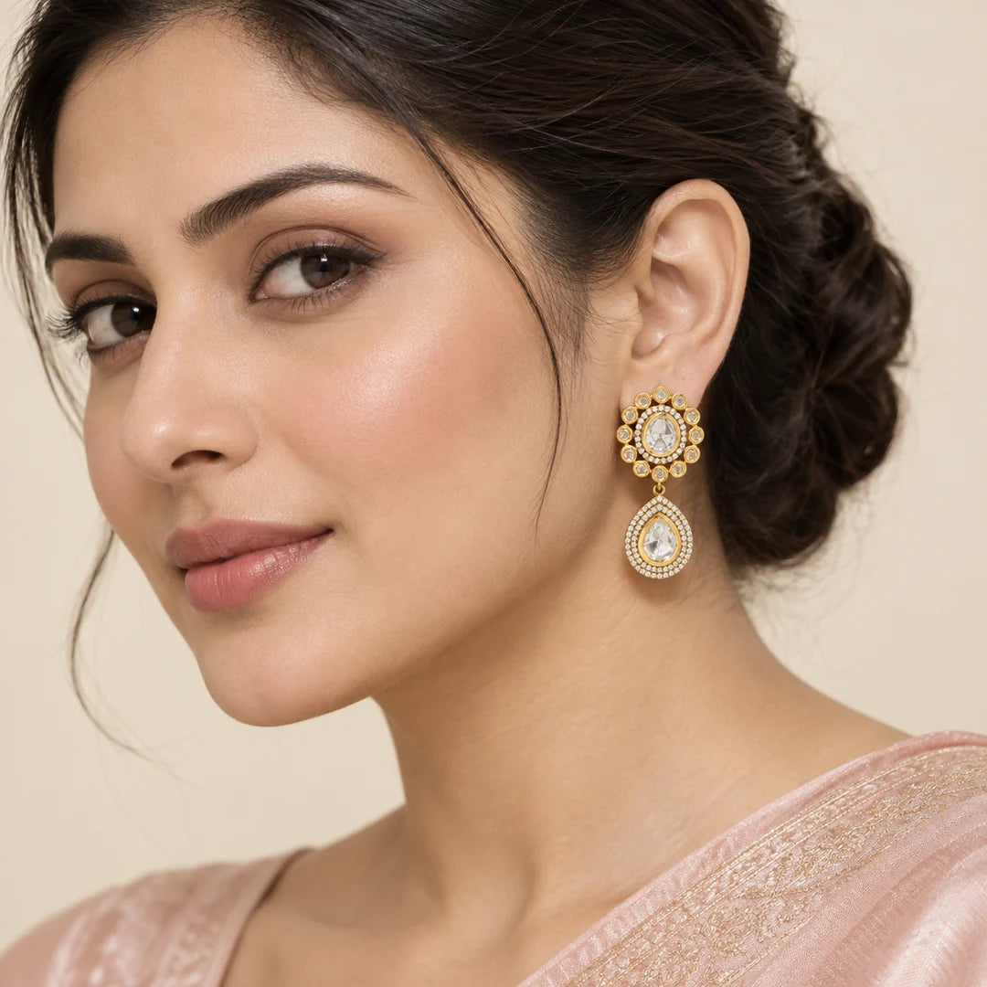 Model in a blush saree wearing Aabha gold-tone earrings with clear stone rosettes and teardrop pendants.