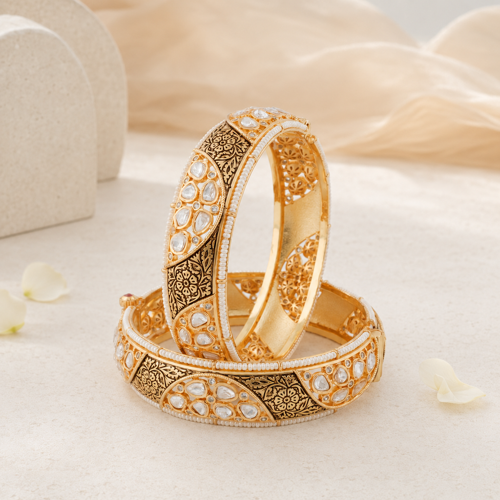 Two ornate gold-toned bangles with intricate filigree and white stone accents, arranged on a soft cream fabric background with blurred decorative props.