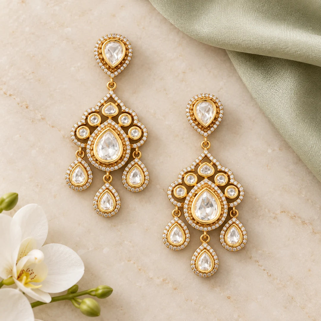 Pair of Advika gold-finish Kundan chandelier earrings on ivory stone beside sage silk and a white orchid.