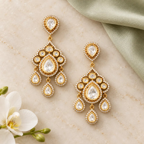 Pair of Advika gold-finish Kundan chandelier earrings on ivory stone beside sage silk and a white orchid.