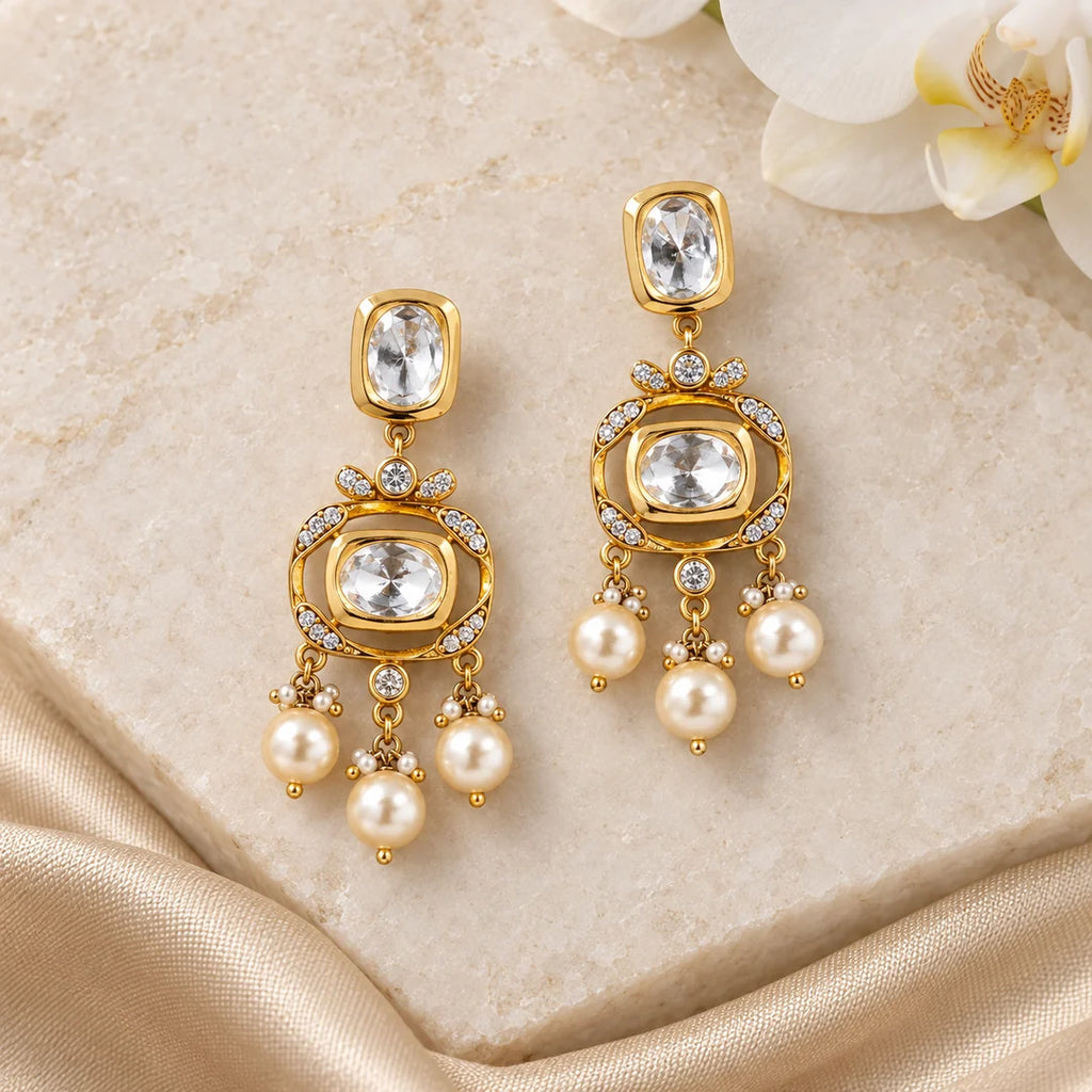Pair of Charvi gold-tone Kundan-style earrings with open frames and cream pearl-style drops on ivory stone beside champagne silk