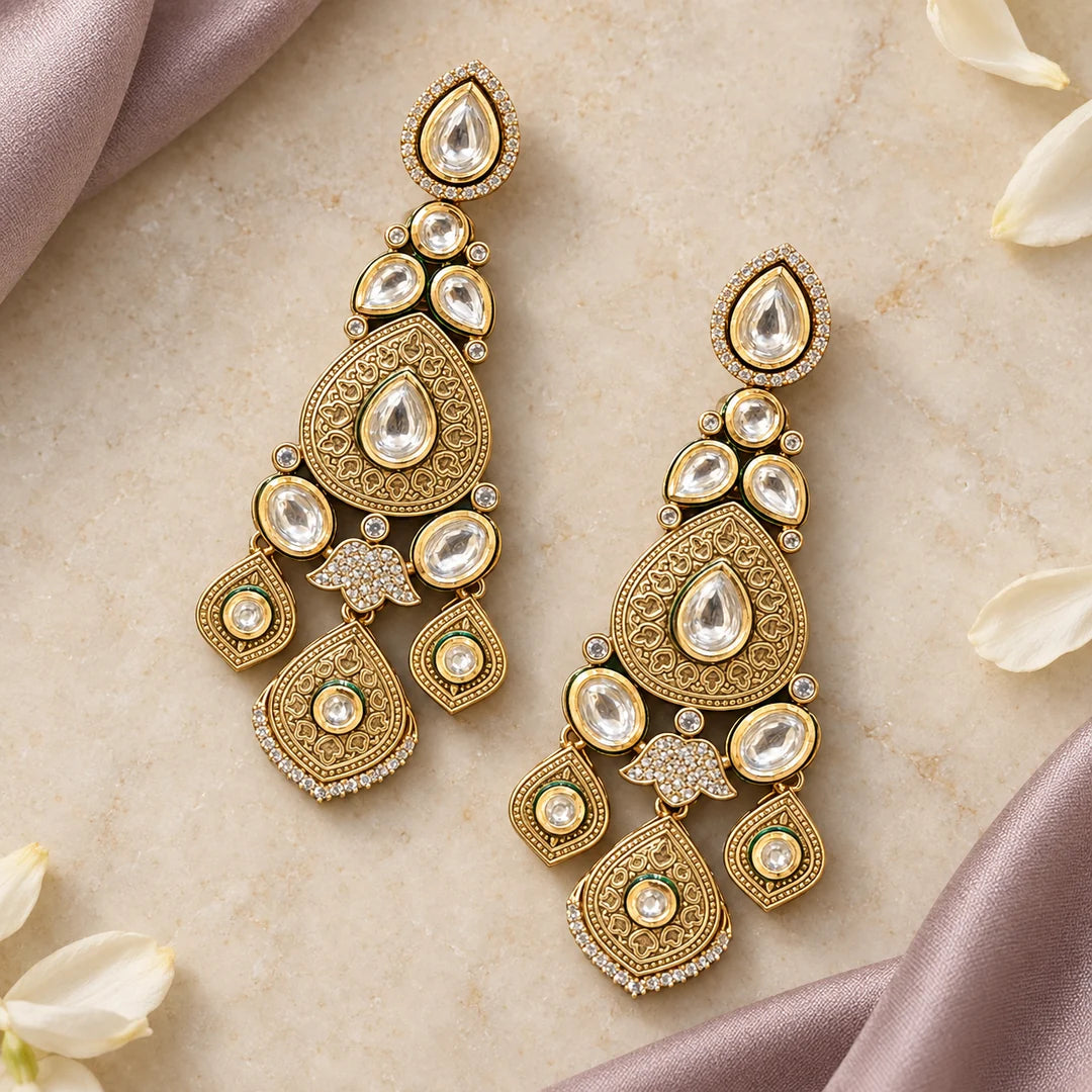 Pair of engraved gold-tone chandelier earrings lying flat on cream stone beside muted mauve silk and ivory petals.