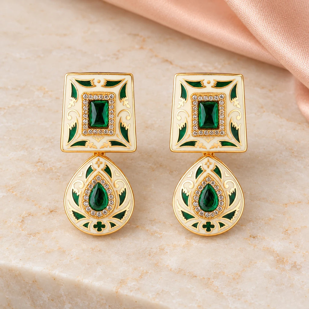 Pair of Kaashni ivory and green drop earrings with gold-tone leaf detailing and clear accents on cream stone beside peach silk.