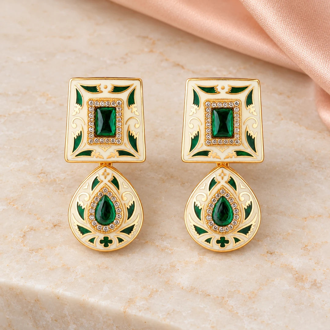 Pair of Kaashni ivory and green drop earrings with gold-tone leaf detailing and clear accents on cream stone beside peach silk.
