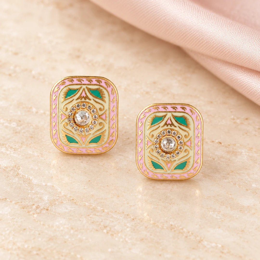 Pair of Kairavi pink and turquoise-colour rectangular stud earrings on cream stone beside blush silk, showing gold-tone botanical detailing.