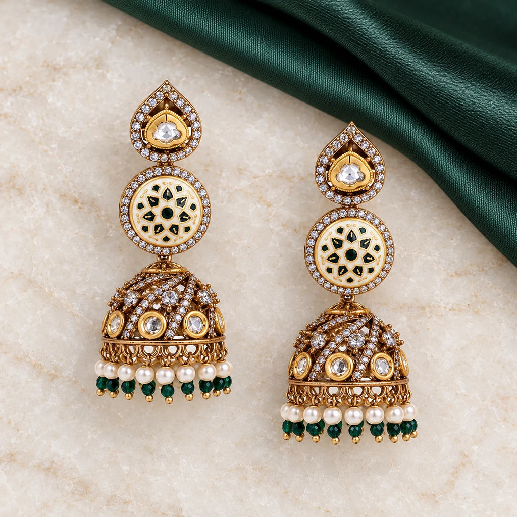 Pair of Kalika ivory and green floral jhumka earrings on cream stone beside forest-green silk, showing stone-set gold-tone domes.