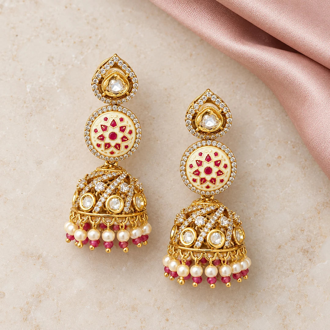Pair of Kalika ivory and ruby-colour floral jhumka earrings on cream stone beside dusty-rose silk, showing stone-set gold-tone domes.