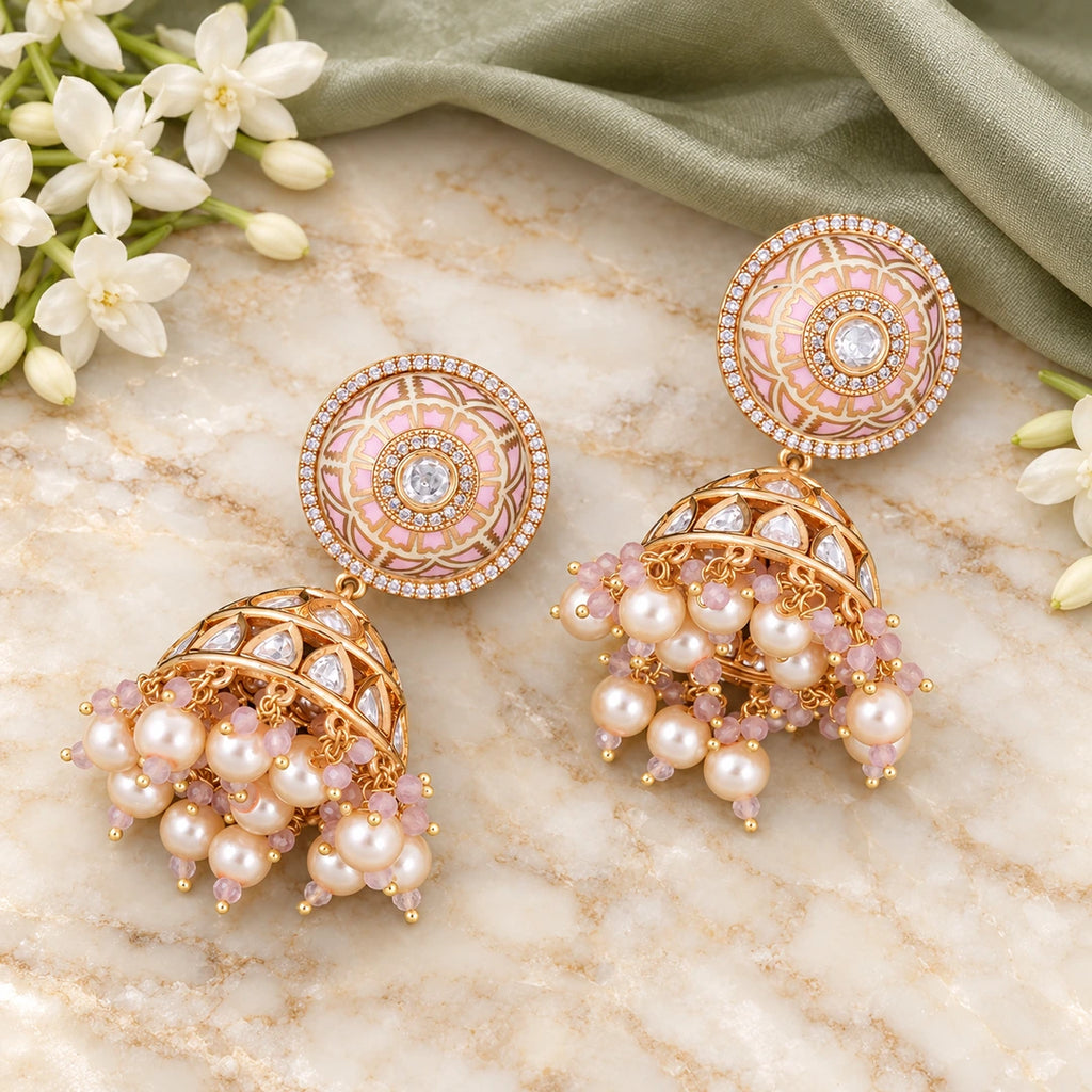 Pair of Kamala ivory and blush jhumka earrings on cream stone beside sage-green silk and white jasmine flowers.
