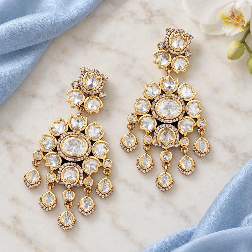 Pair of clear stone floral chandelier earrings lying flat on ivory marble beside pale blue silk and a white orchid.