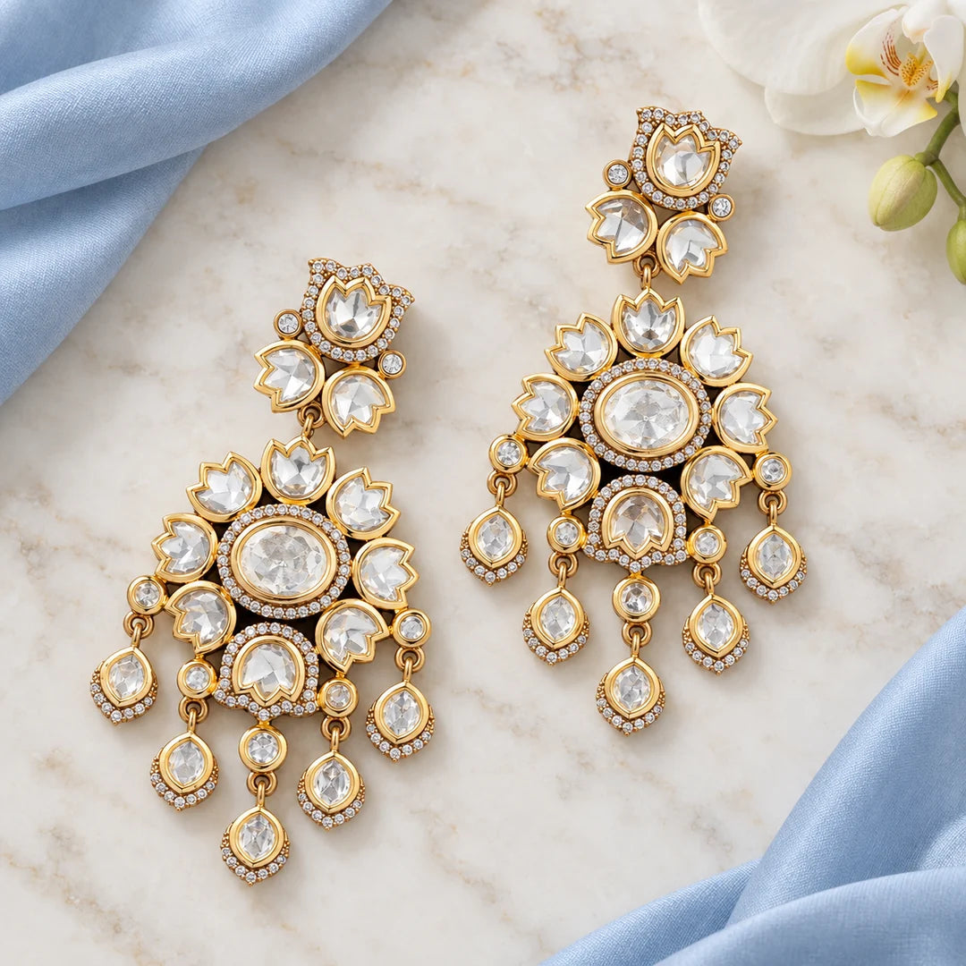 Pair of clear stone floral chandelier earrings lying flat on ivory marble beside pale blue silk and a white orchid.