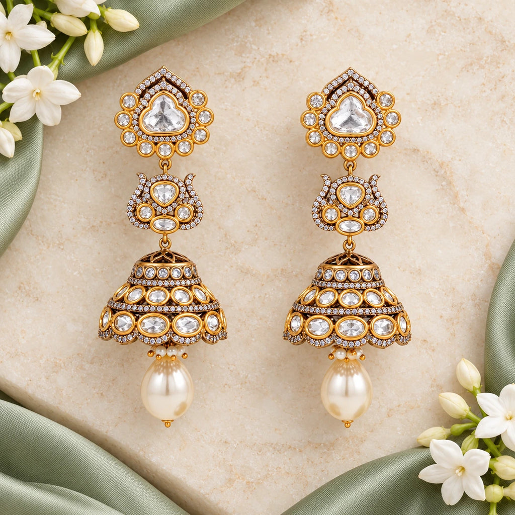 Pair of Kamya clear and pearl-style jhumka earrings on cream stone beside sage silk and white jasmine flowers.