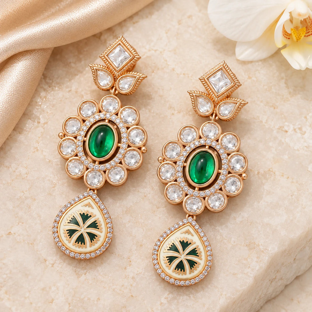 Pair of Kamya ivory and green floral drop earrings on cream stone beside champagne silk and a white orchid.