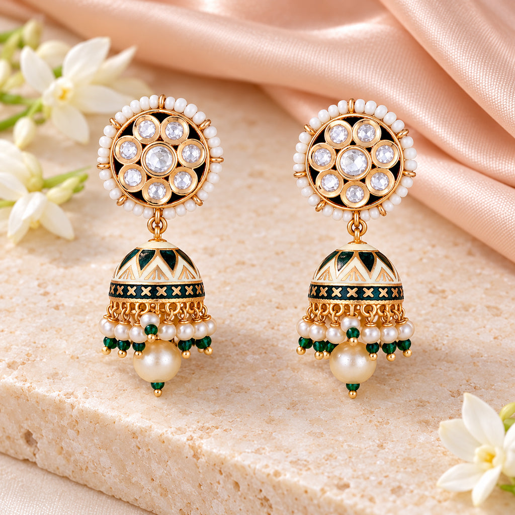 Pair of Kanavi ivory and green pearl-style jhumka earrings on cream stone beside peach silk and white flowers.