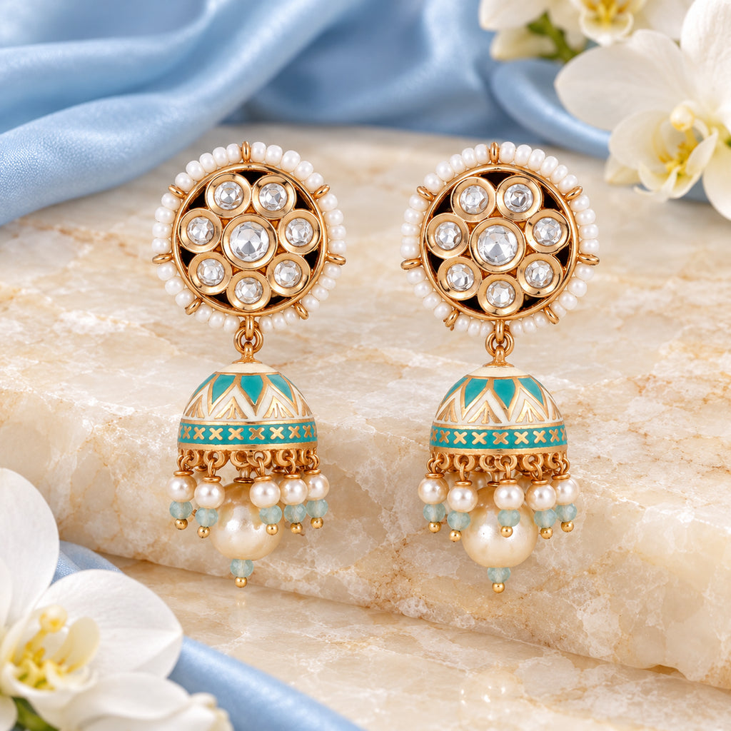 Pair of Kanavi ivory and turquoise pearl-style jhumka earrings on cream stone beside powder blue silk and white orchids.