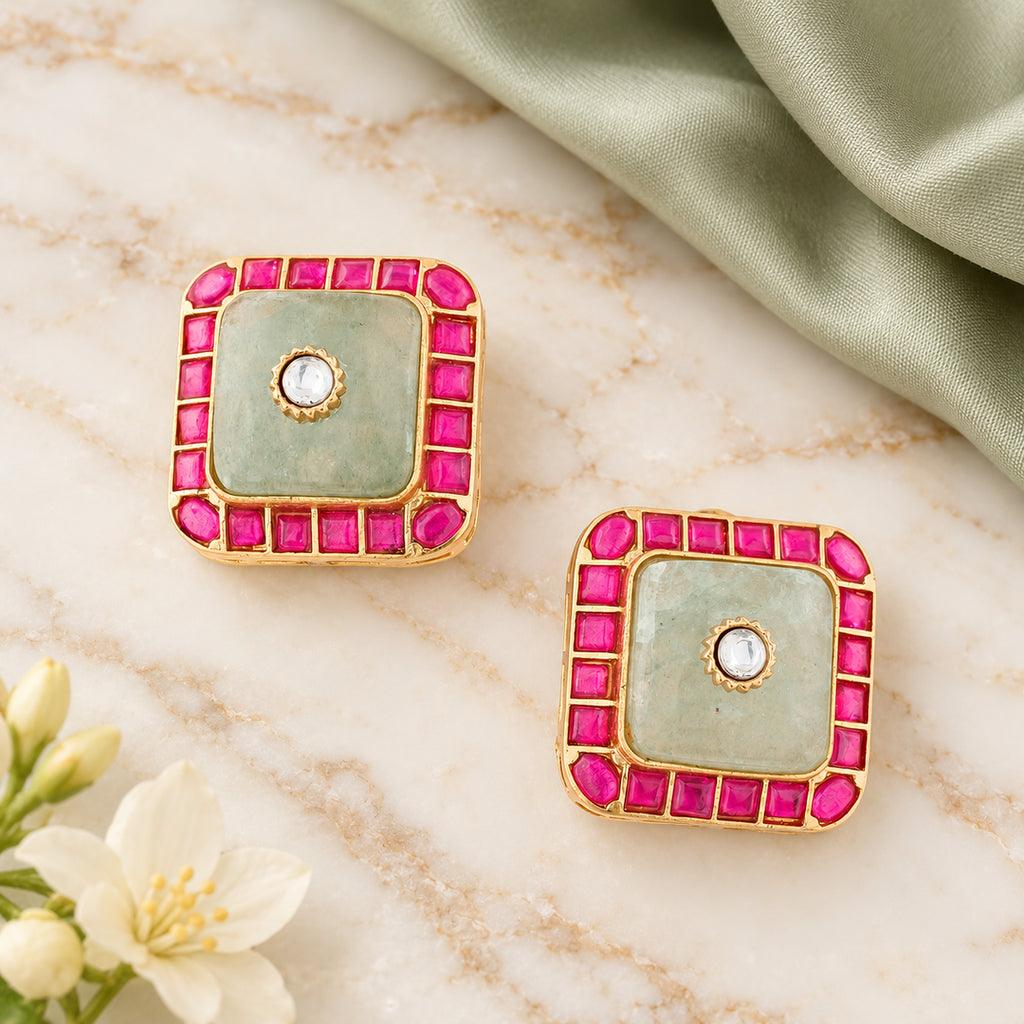Pair of Kashika sage green and ruby-colour square stud earrings on cream marble beside sage silk and cream flowers.
