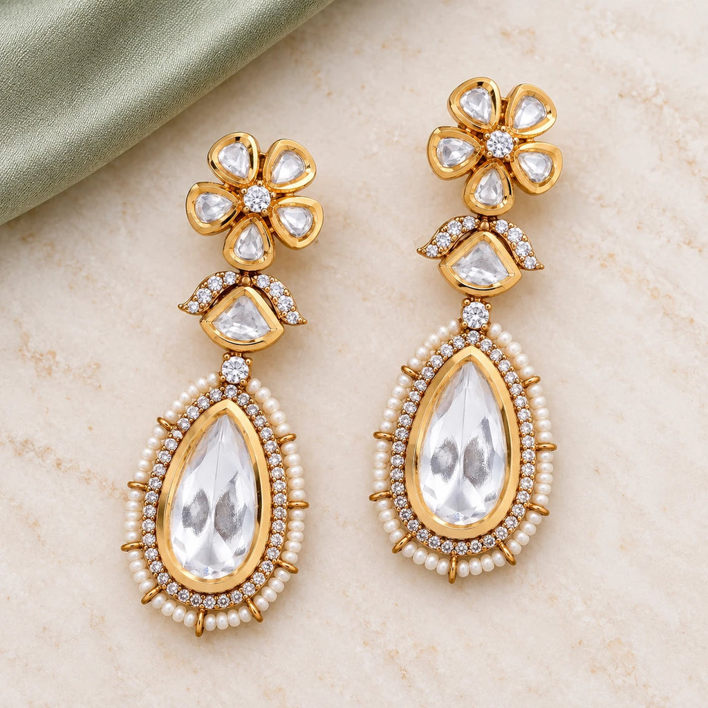 Pair of Kashmira clear and pearl-style floral teardrop earrings on pale cream stone beside sage fabric.