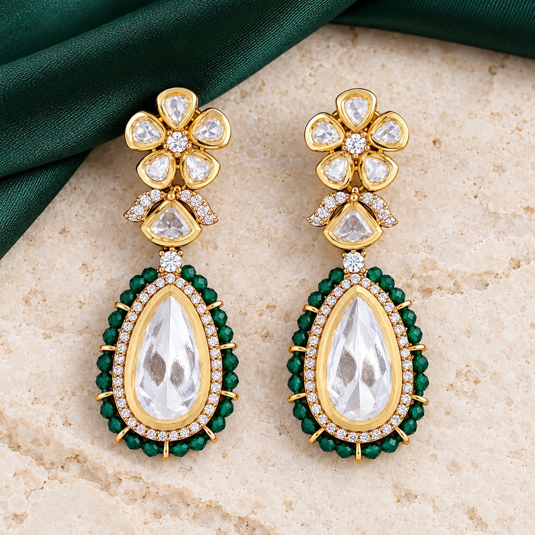 Pair of Kashmira green floral teardrop earrings with clear centres and gold-tone settings on cream stone beside green fabric.