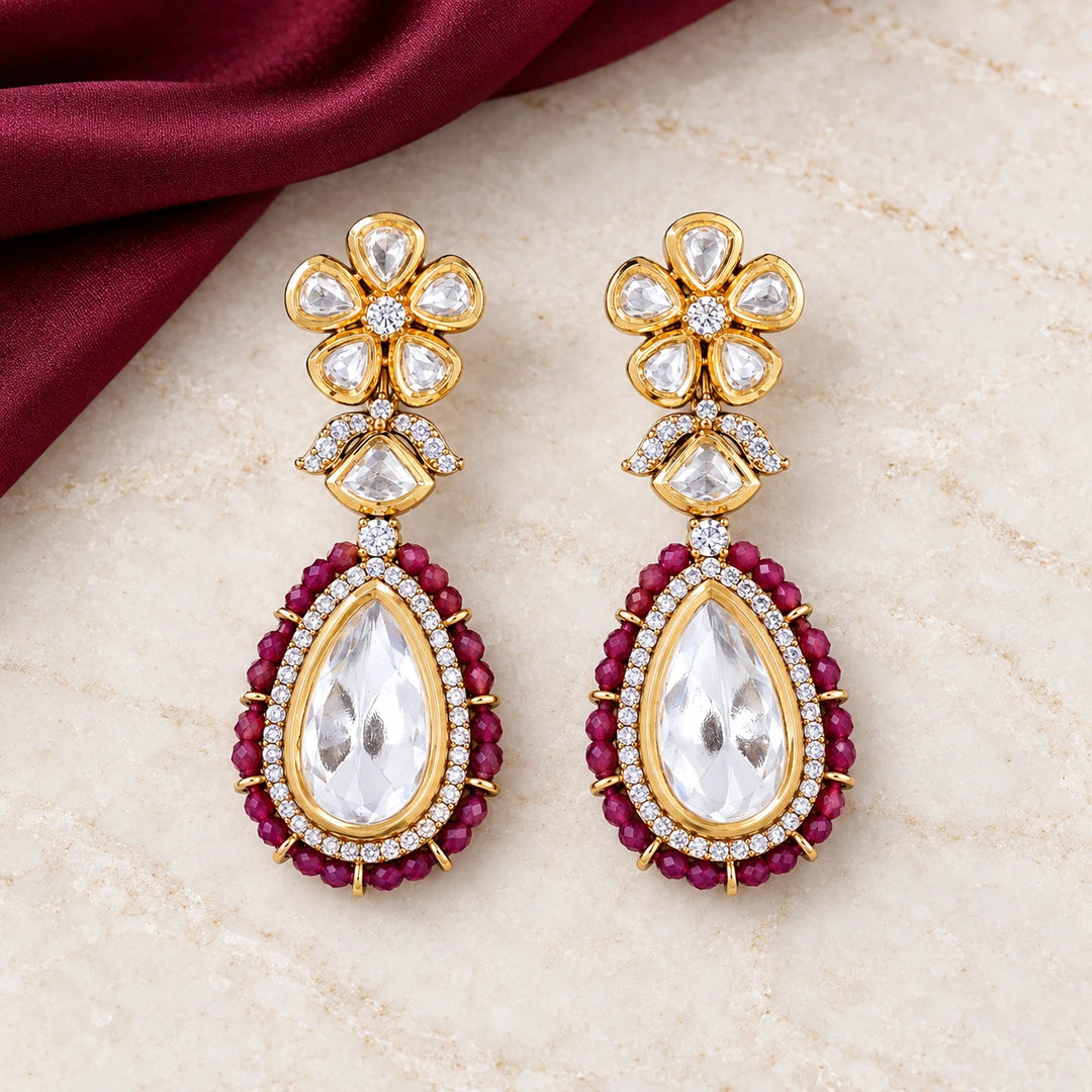 Pair of Kashmira ruby-colour floral teardrop earrings with clear stones and gold-tone settings on ivory stone beside burgundy fabric.
