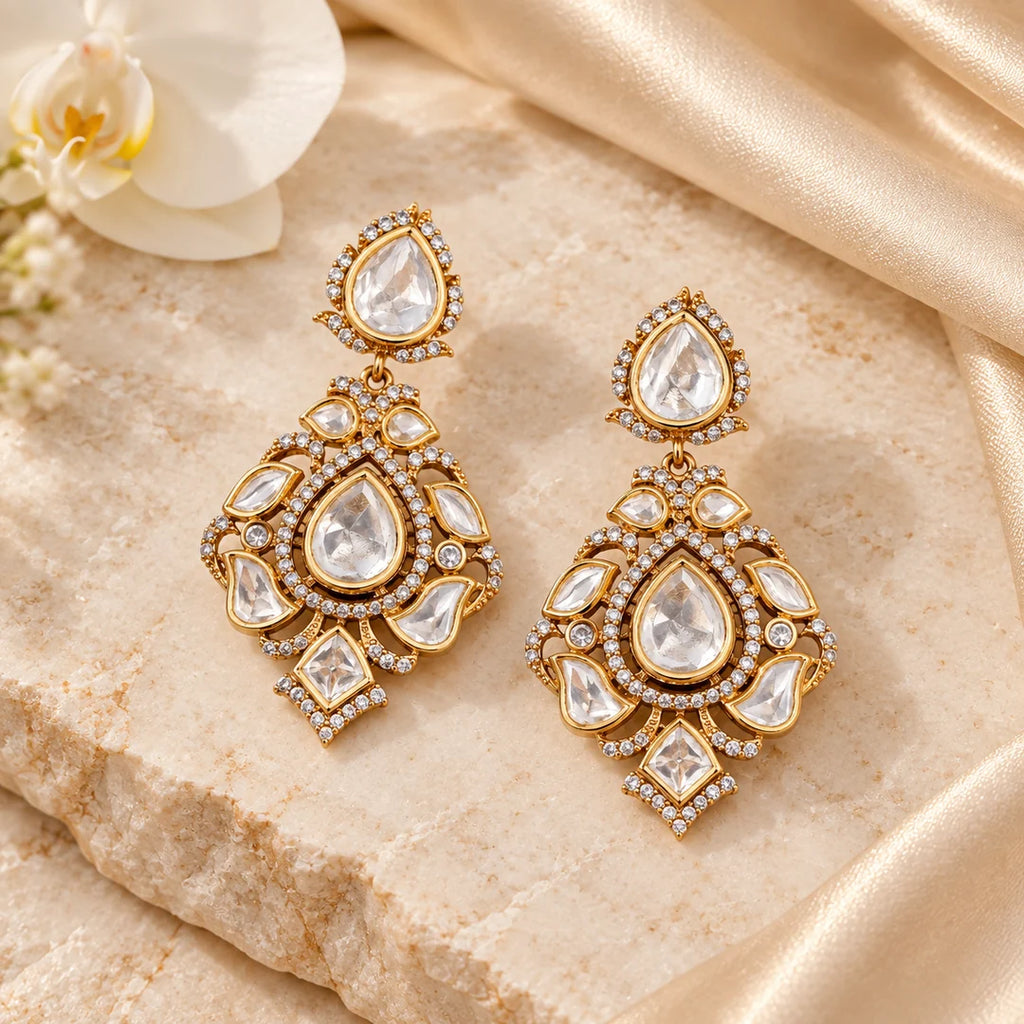 Pair of Kashvi gold-tone floral drop earrings with pear-shaped clear stones arranged on ivory stone beside champagne silk and a white orchid.