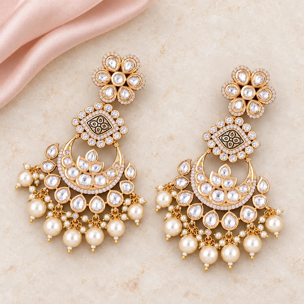 Pair of Kaumudi clear-stone chandbali earrings with patterned gold-tone connectors and cream pearl-style drops on cream stone beside blush silk.