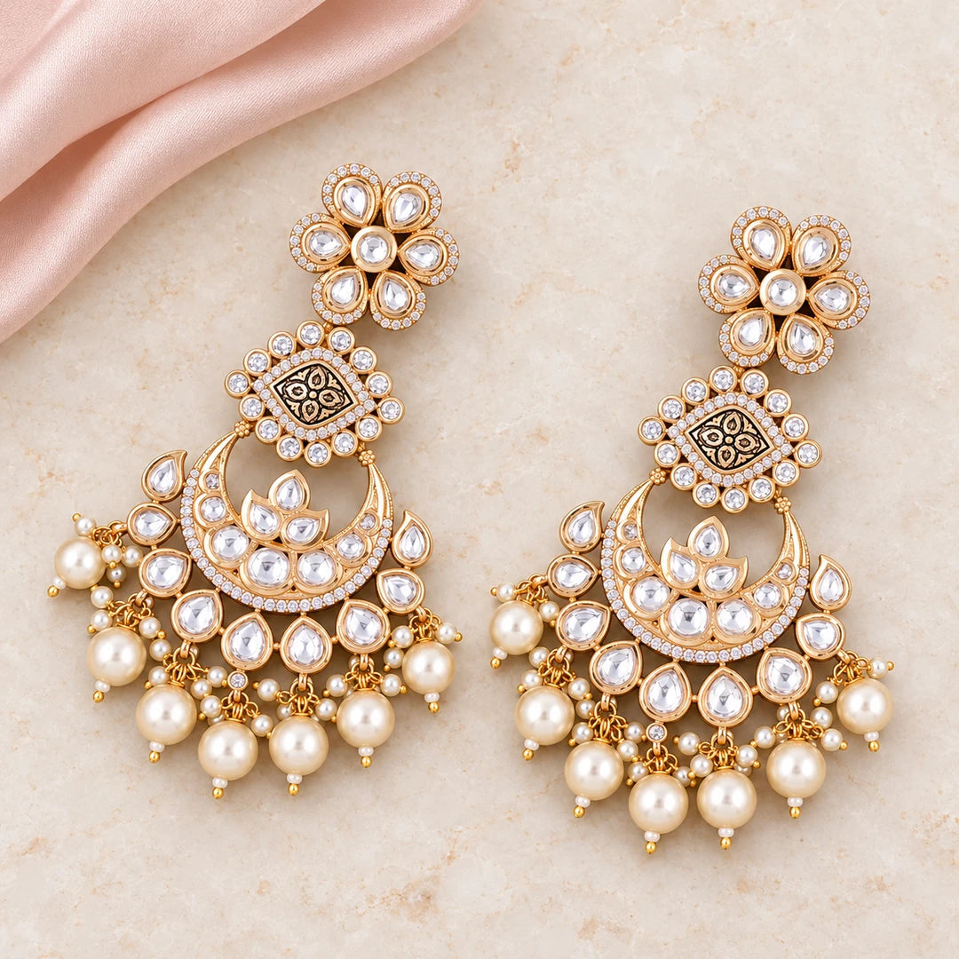 Pair of Kaumudi clear-stone chandbali earrings with patterned gold-tone connectors and cream pearl-style drops on cream stone beside blush silk.
