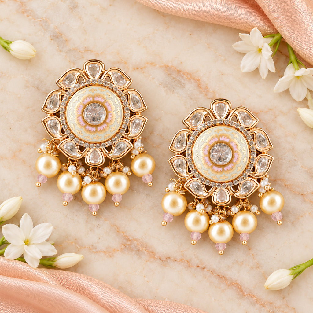 Pair of Kavini ivory and blush pearl-style earrings on cream marble beside peach silk and white jasmine flowers.