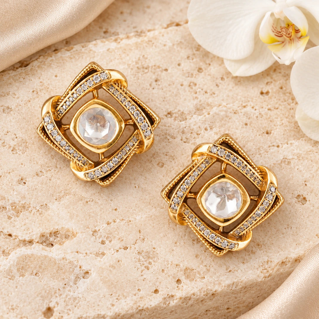 Pair of Kavira clear geometric stud earrings on cream stone beside champagne silk and a white orchid.