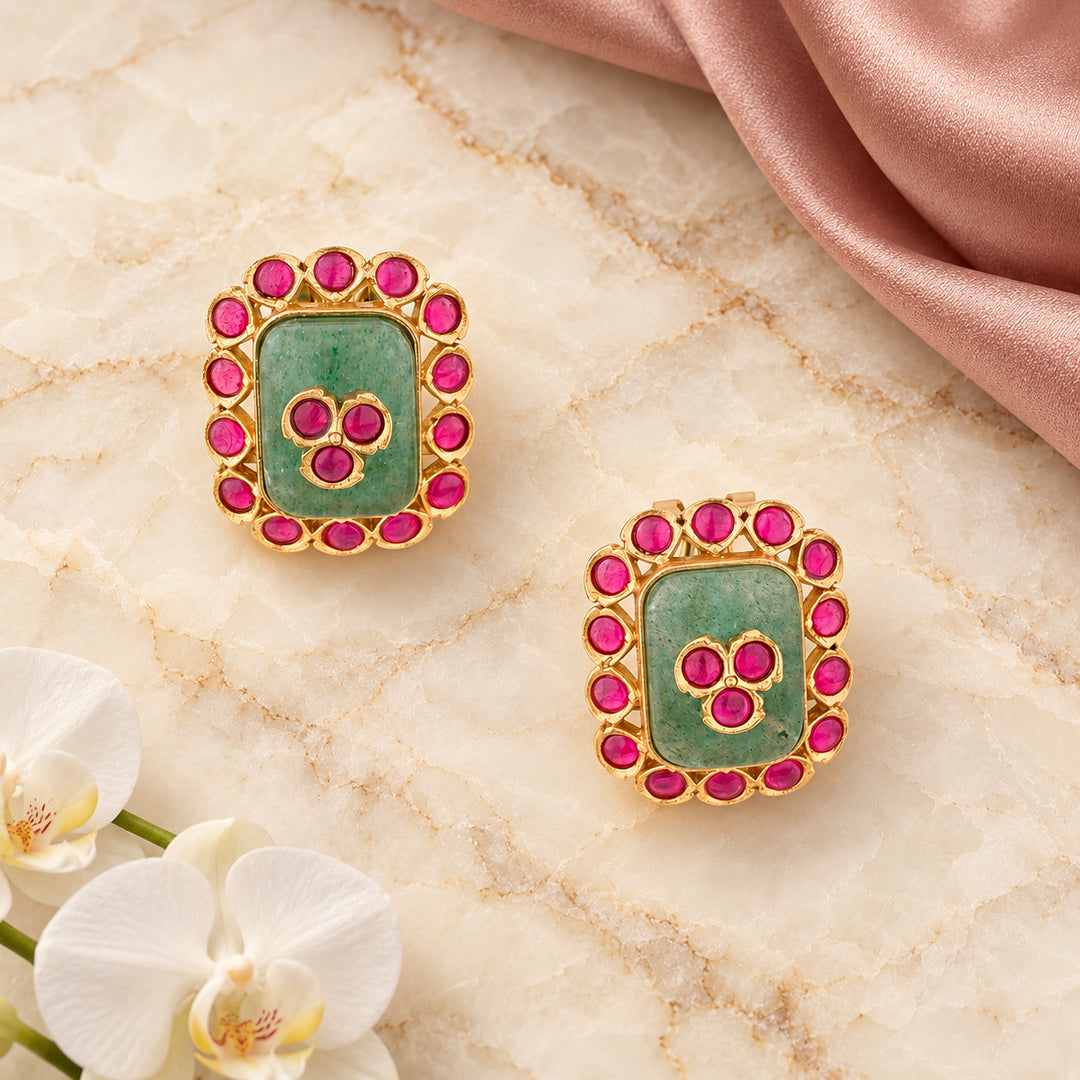 Pair of Kavisha sage green and ruby-colour stud earrings on cream stone beside dusty rose silk and white orchids.