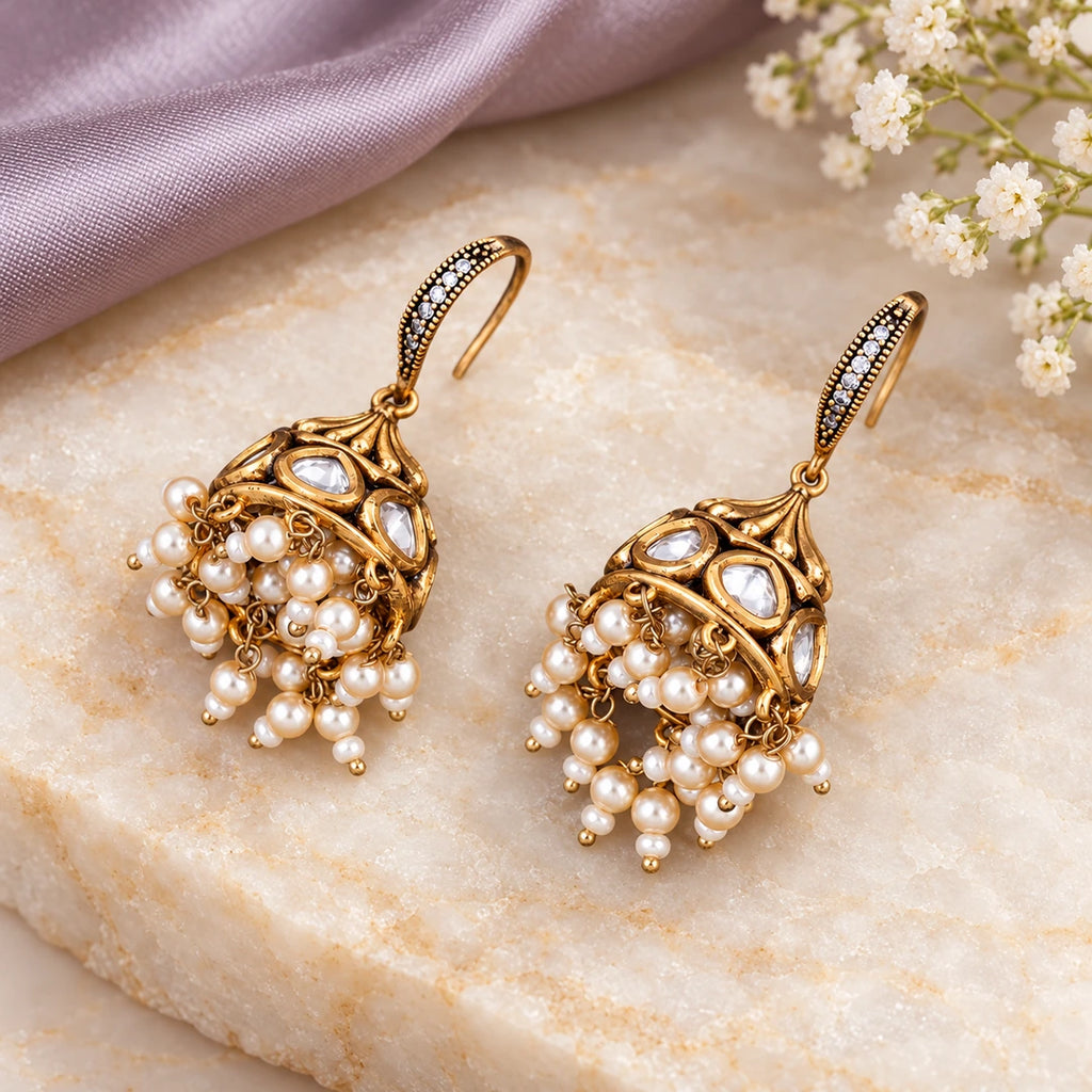 Pair of Kavitha clear and pearl-style hook jhumkas on cream stone beside lavender silk and small white flowers.