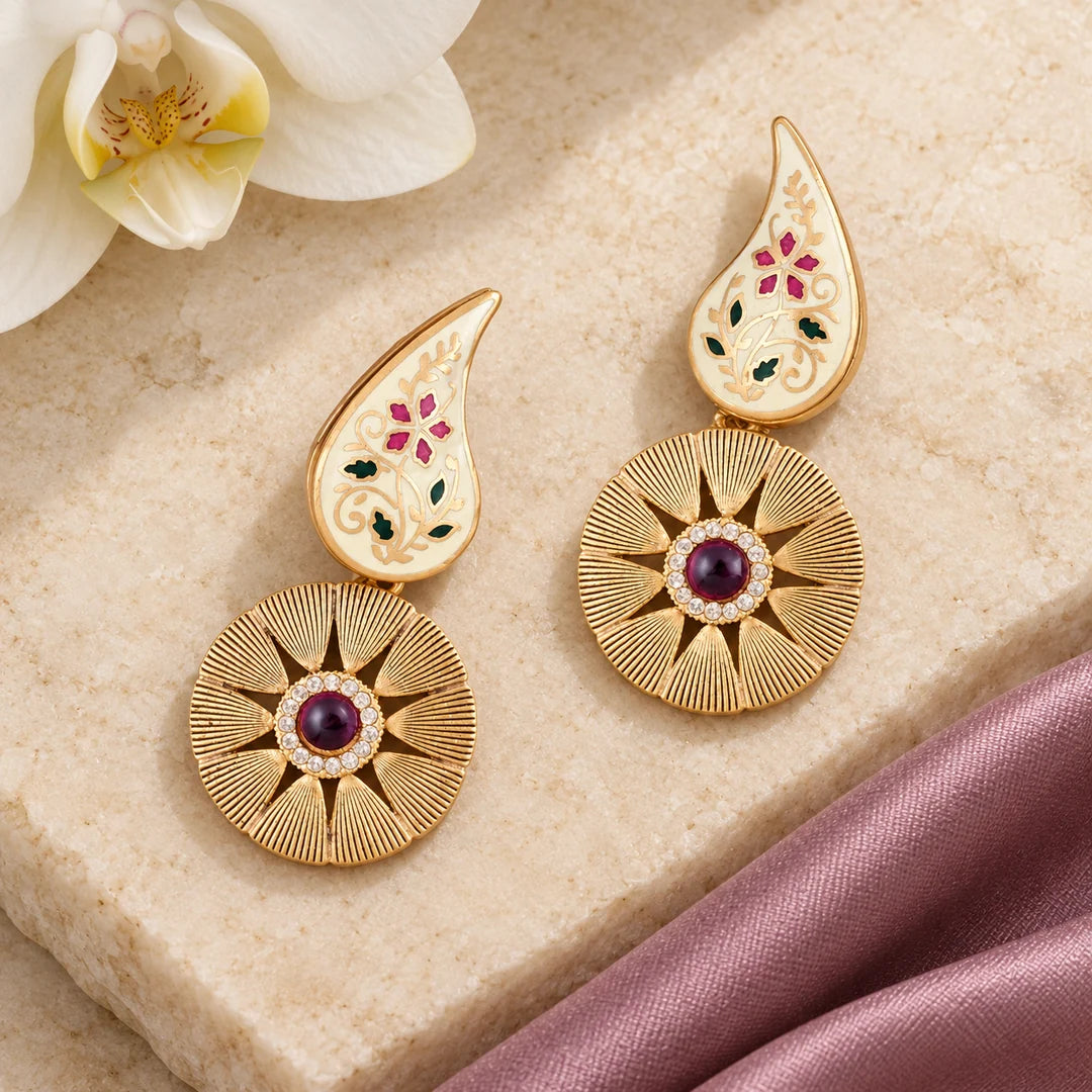 Pair of Kavya ivory and ruby-colour floral drop earrings on cream stone beside mauve silk and a white orchid.