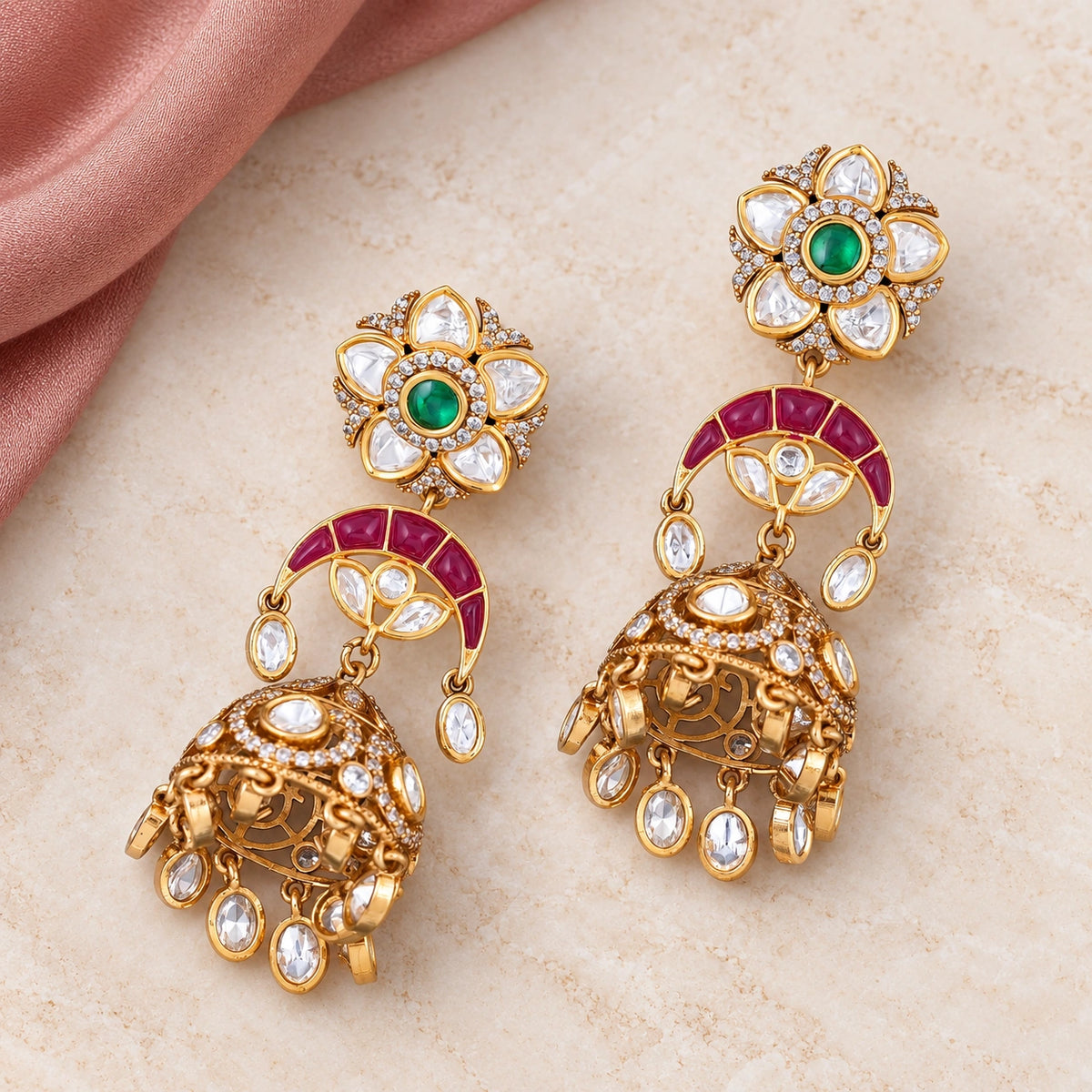 Pair of Keshavi ruby-colour and green floral jhumka earrings with clear stone drops on cream stone beside dusty rose fabric.