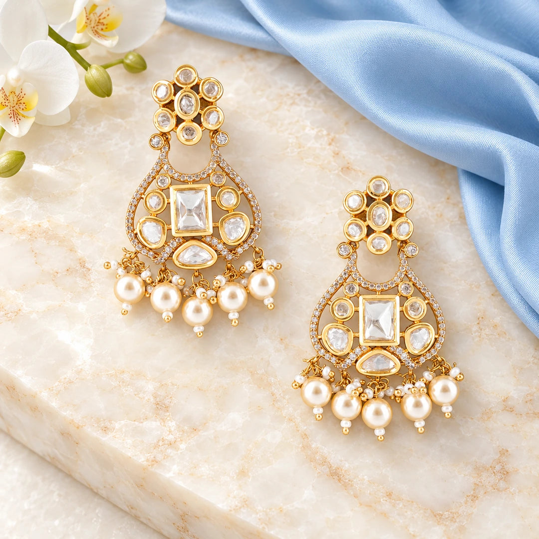 Pair of Ketaki clear and pearl-style chandelier earrings on cream stone beside powder-blue silk and white orchids.
