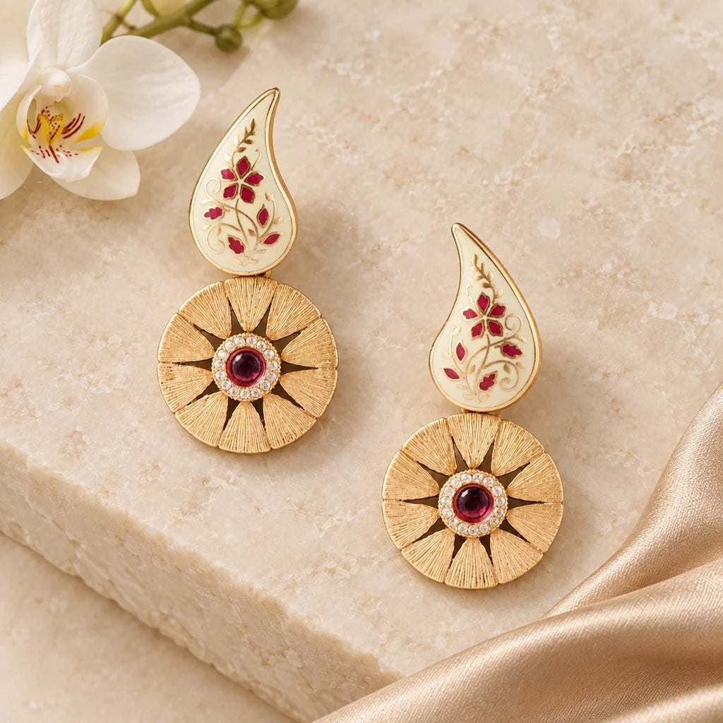 Pair of Kiran ivory and ruby-colour floral drop earrings on cream stone beside champagne silk and a white orchid.