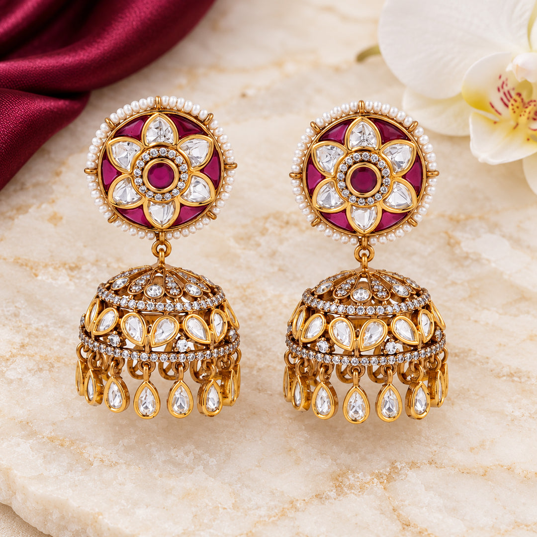 Pair of Kiranya ruby-colour floral jhumka earrings on cream stone beside burgundy silk and a white orchid.