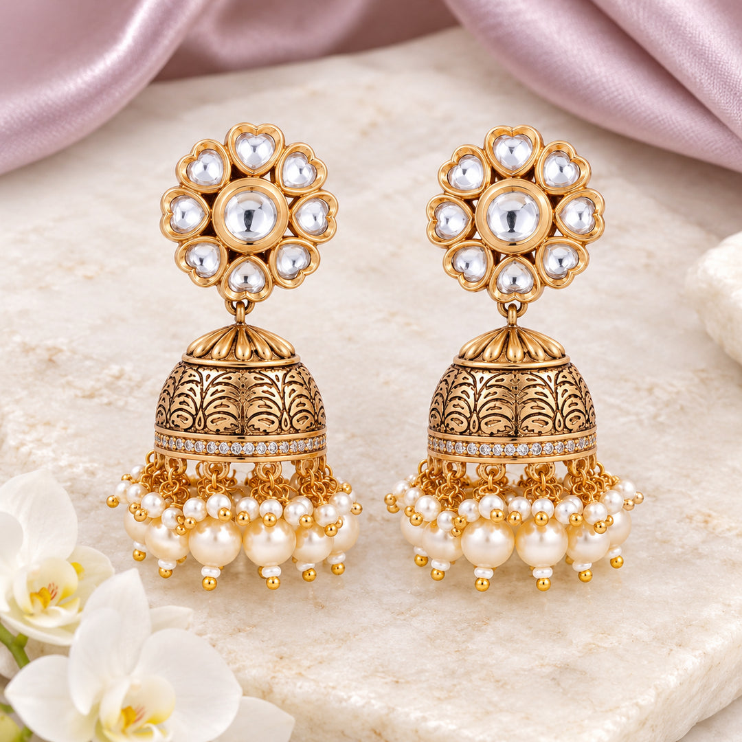 Pair of Kritika clear floral jhumka earrings with engraved gold-tone domes and cream pearl-style drops on cream stone beside mauve silk and a white orchid.