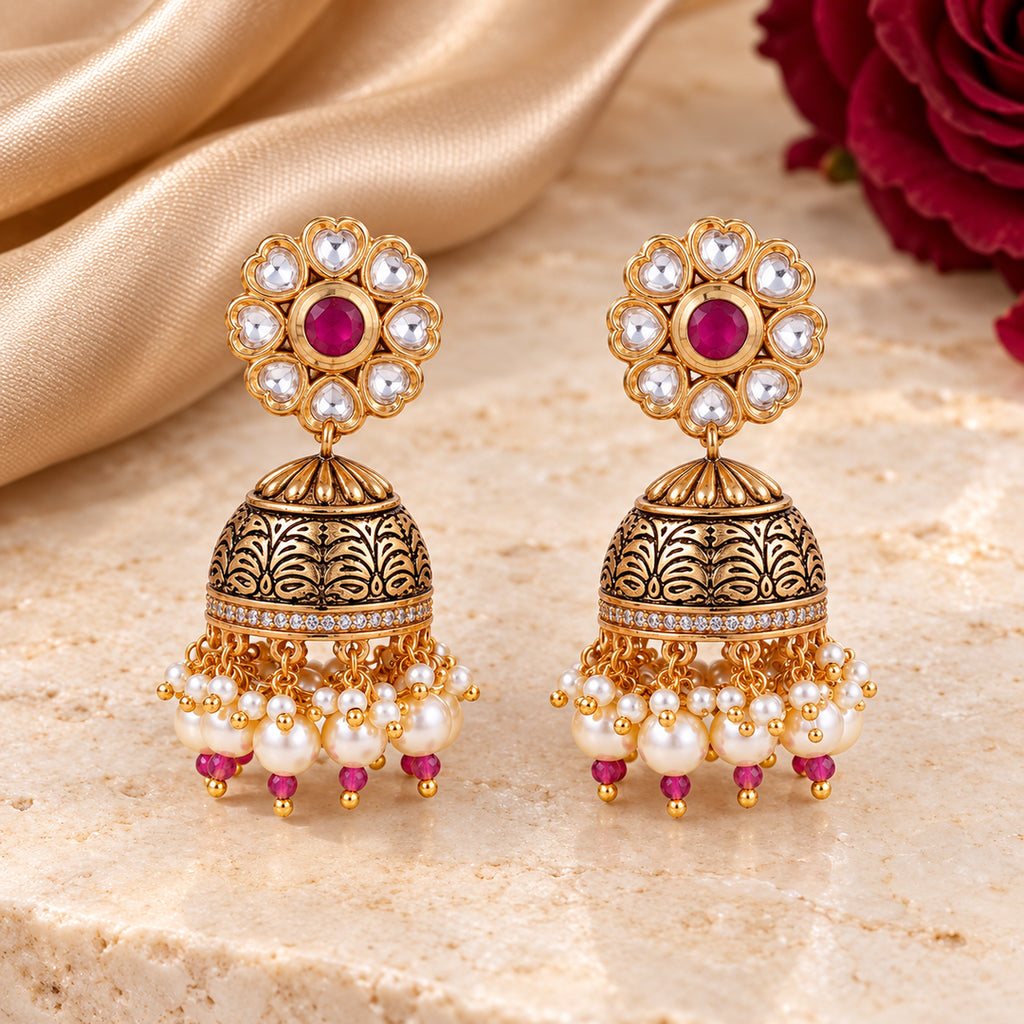 Pair of Kritika ruby-colour floral jhumka earrings with patterned gold-tone domes and pearl-style fringe on cream stone beside champagne silk and a burgundy rose.