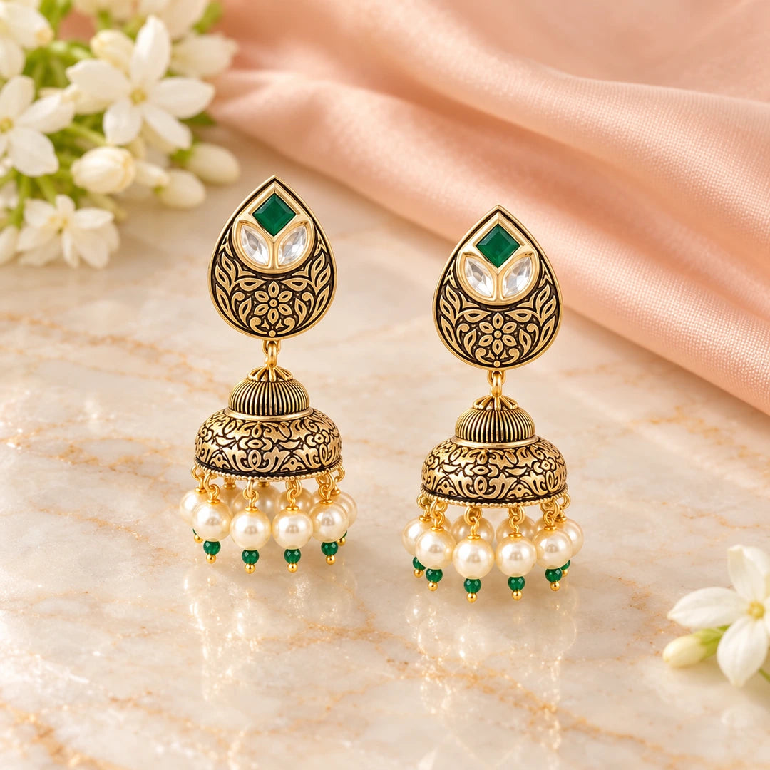 Pair of Kritila green and pearl-style jhumka earrings on cream stone beside peach silk and white jasmine flowers.