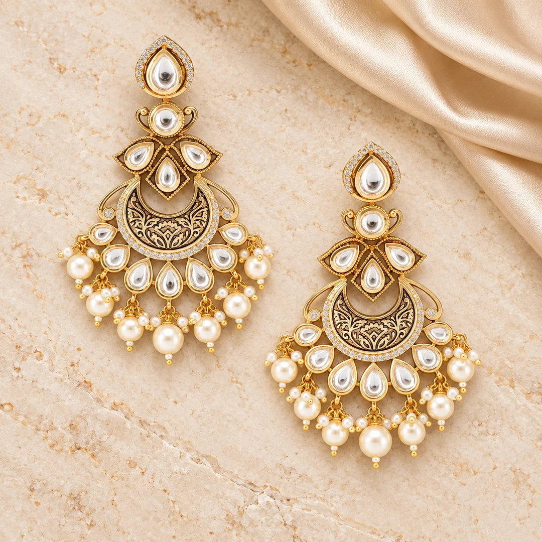 Pair of Kumudini clear and pearl-style chandbali earrings on cream stone beside champagne silk, showing engraved gold-tone crescents.