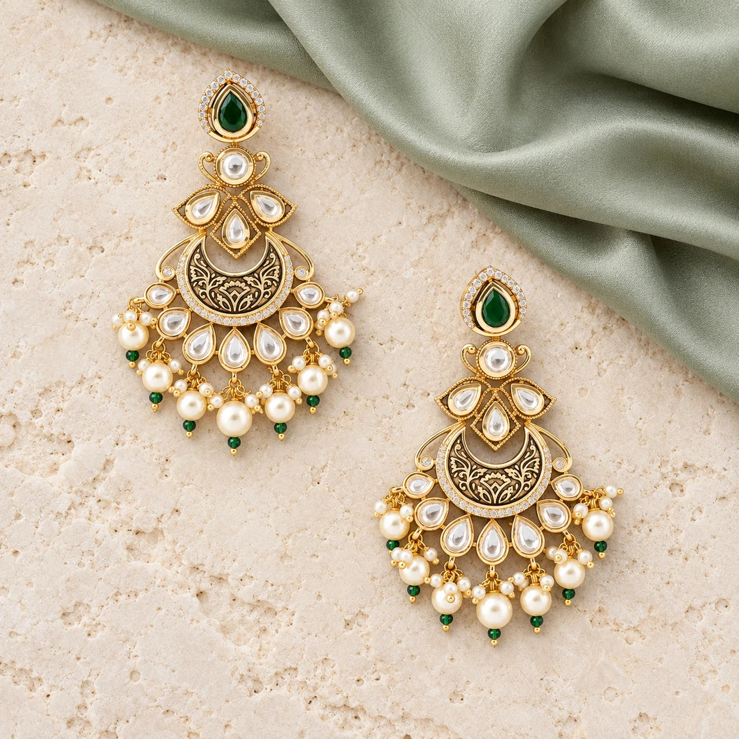 Pair of Kumudini green and pearl-style chandbali earrings on cream stone beside sage silk, showing clear stones and engraved gold-tone crescents.